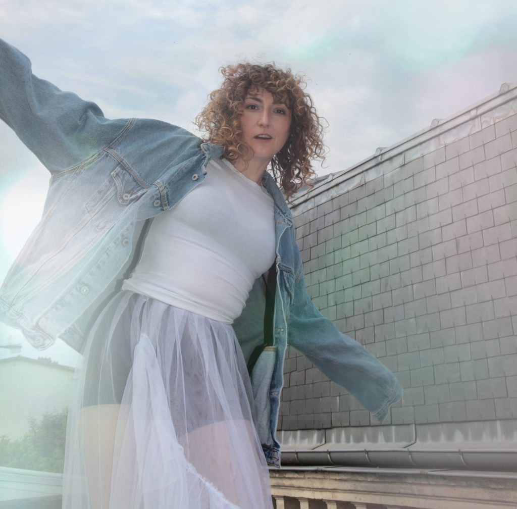 Une femme aux cheveux bouclés porte une veste en jean et une jupe en tulle, se tenant debout sur un toit sous un ciel nuageux.